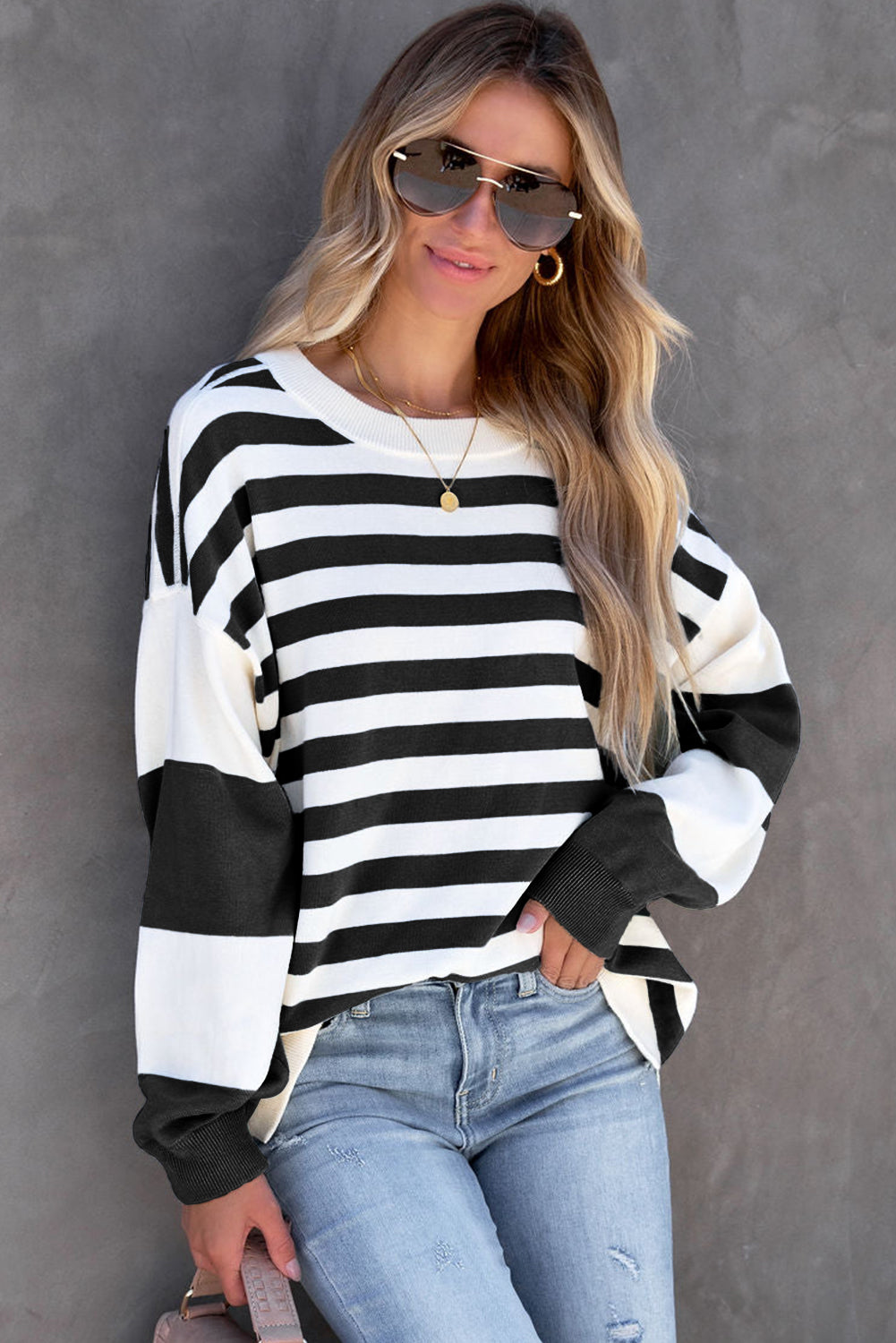 A person stands against a gray background, wearing sunglasses, a relaxed-fit Black Stripe Drop Shoulder Striped Pullover Sweatshirt, and ripped blue jeans, with long, wavy blonde hair.