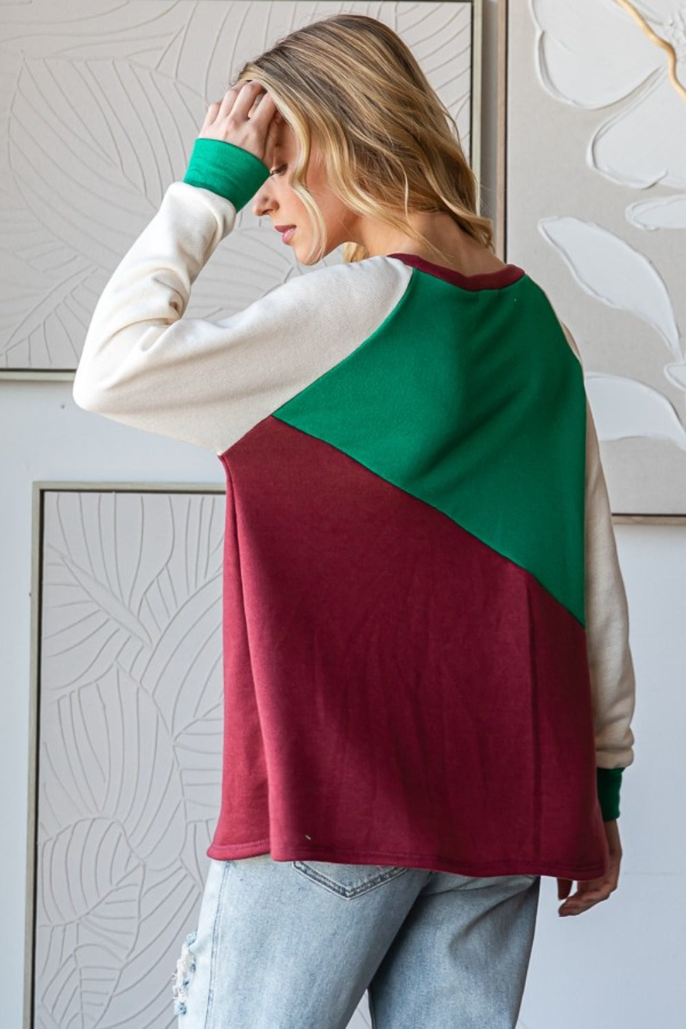 A person with long, wavy hair is wearing a HOPELY Full Size Color Block Long Sleeve T-Shirt in green, red, and white paired with light blue jeans, standing indoors against a patterned wall.