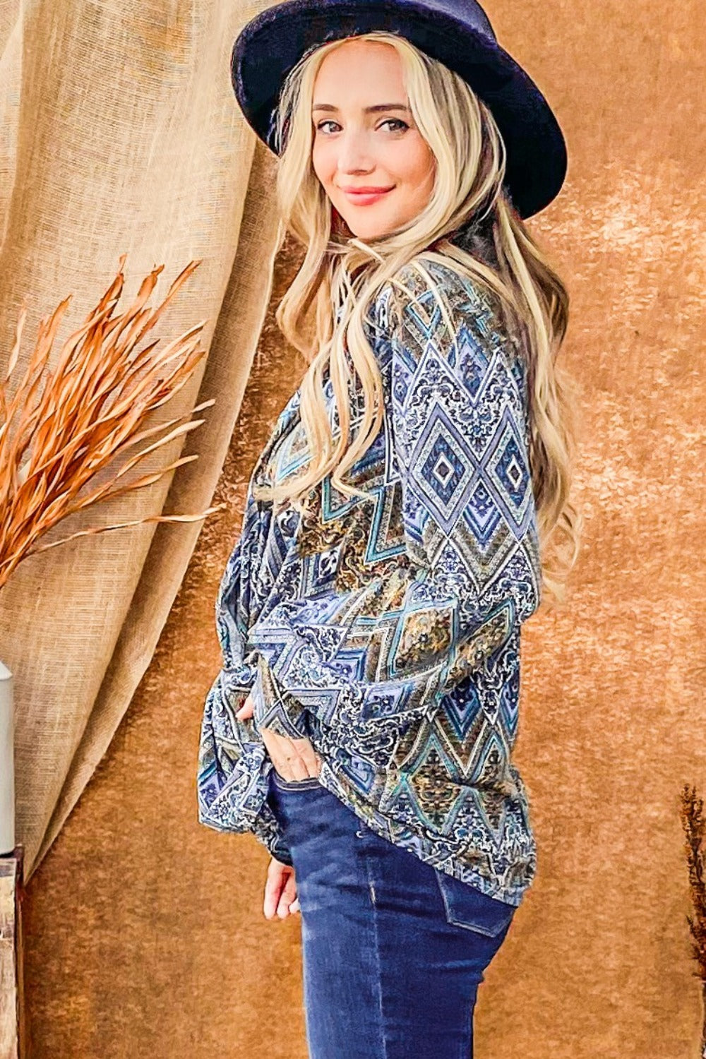 A woman with long blonde hair wearing an And The Why Print Twist Knot Long Sleeve Blouse, blue jeans, and a dark blue hat stands in front of a brown backdrop.