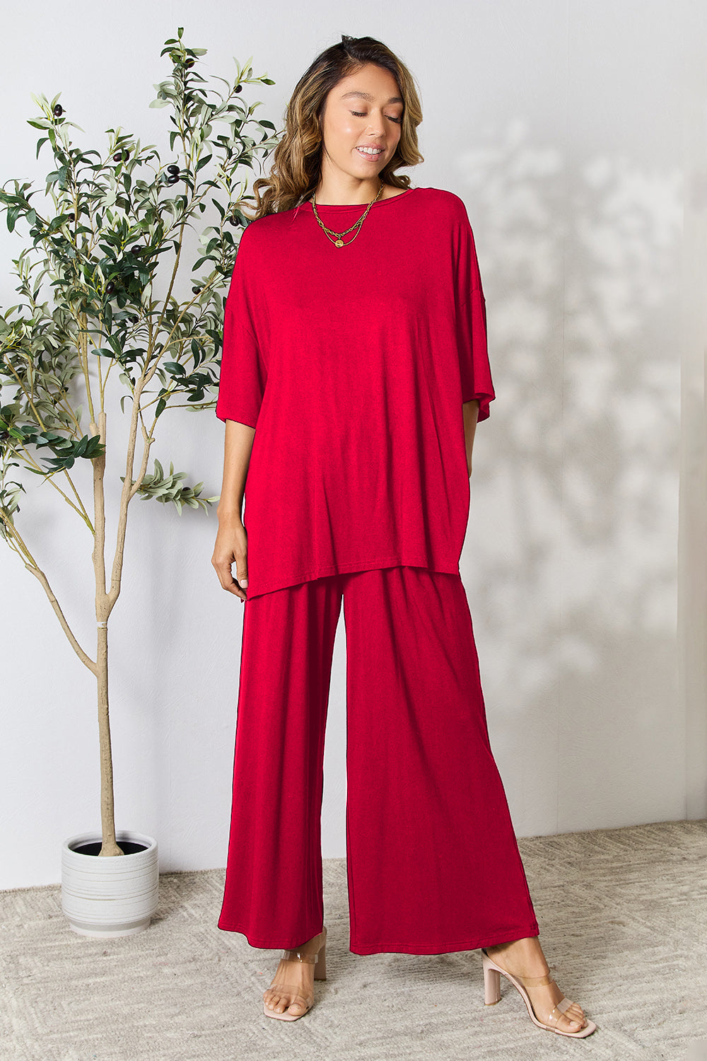 A woman stands near a potted plant and wooden ladder, wearing the Double Take Full Size Round Neck Slit Top and Pants Set in a bright pink two-piece outfit with matching wide-leg pants that are available in plus size and are easily machine washable.