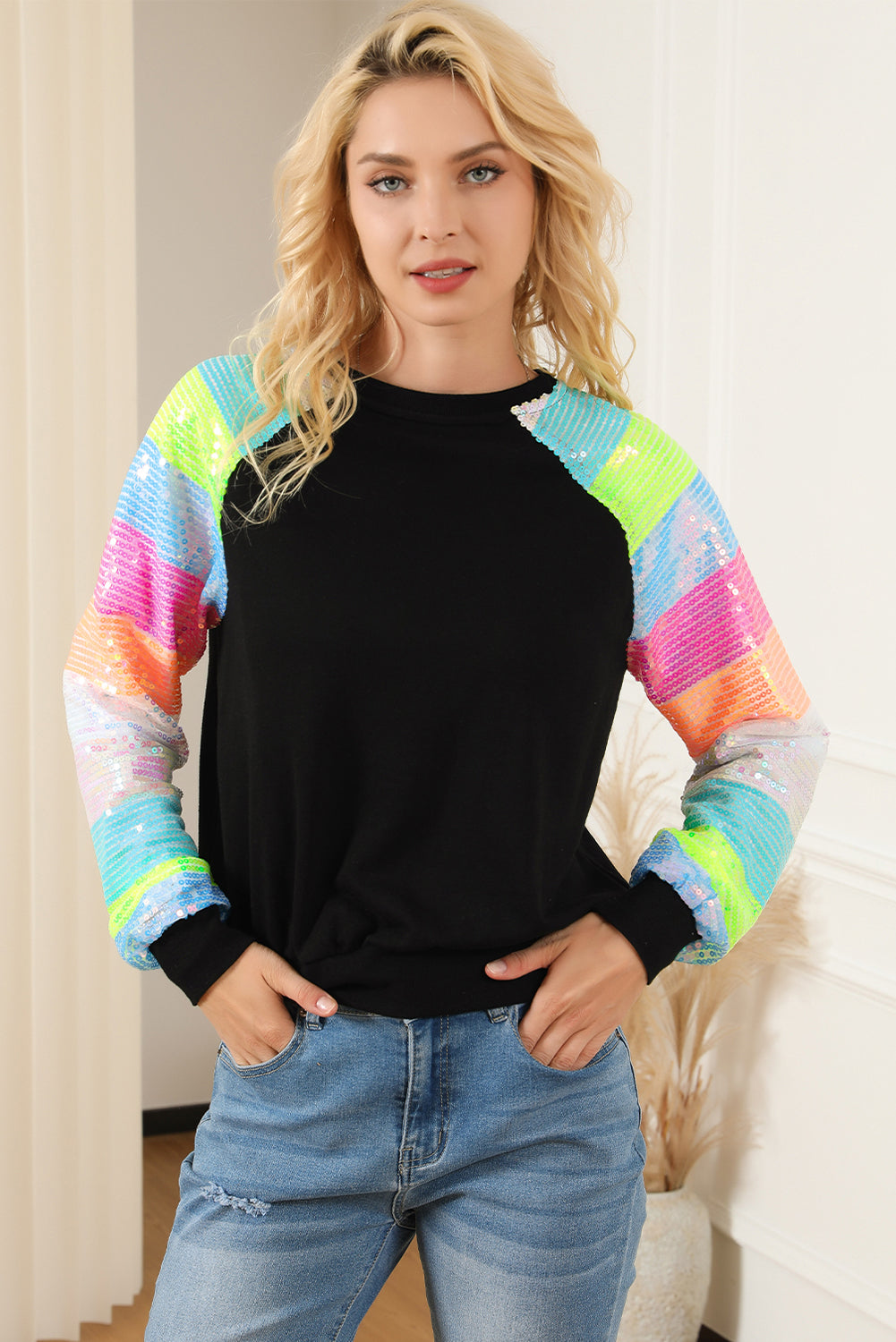 A person with shoulder-length hair is wearing a Black Sequin Color Block Raglan Sleeve Pullover Sweatshirt and blue jeans. They are facing away from the camera in a room with a potted plant.