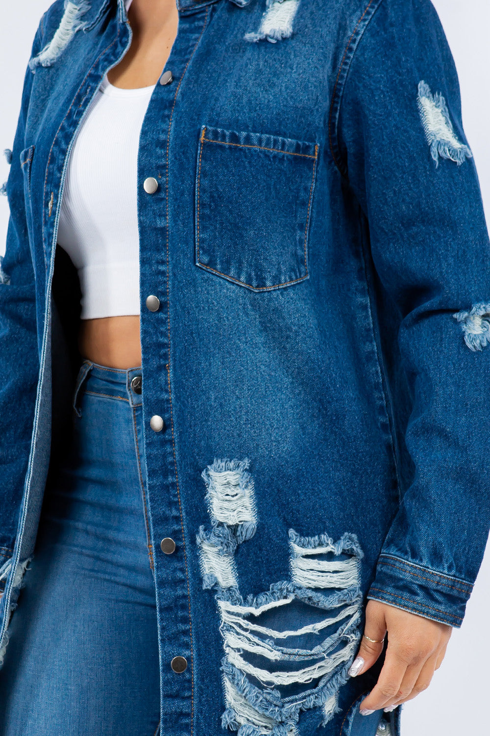 A woman is wearing an American Bazi Distressed Button Up Long Sleeve Denim Jacket over a white top and distressed jeans, posing against a plain background. The denim jacket adds an edgy touch to her chic ensemble.