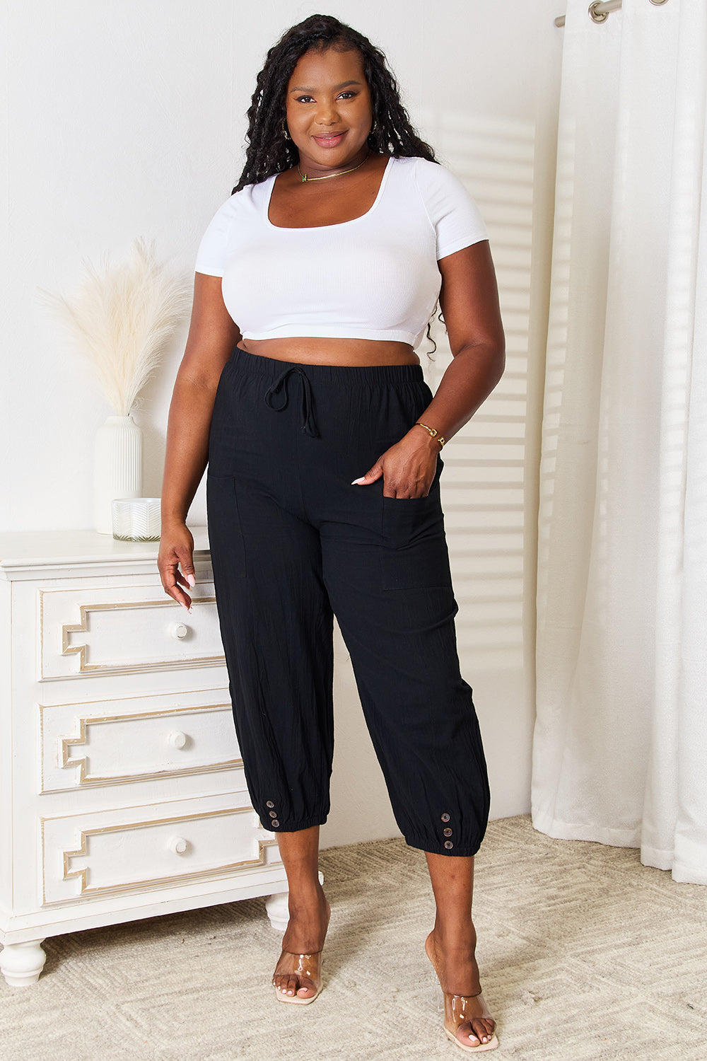 A person wearing Shiny Decorative Button Cropped Pants and a white top stands near a white dresser.