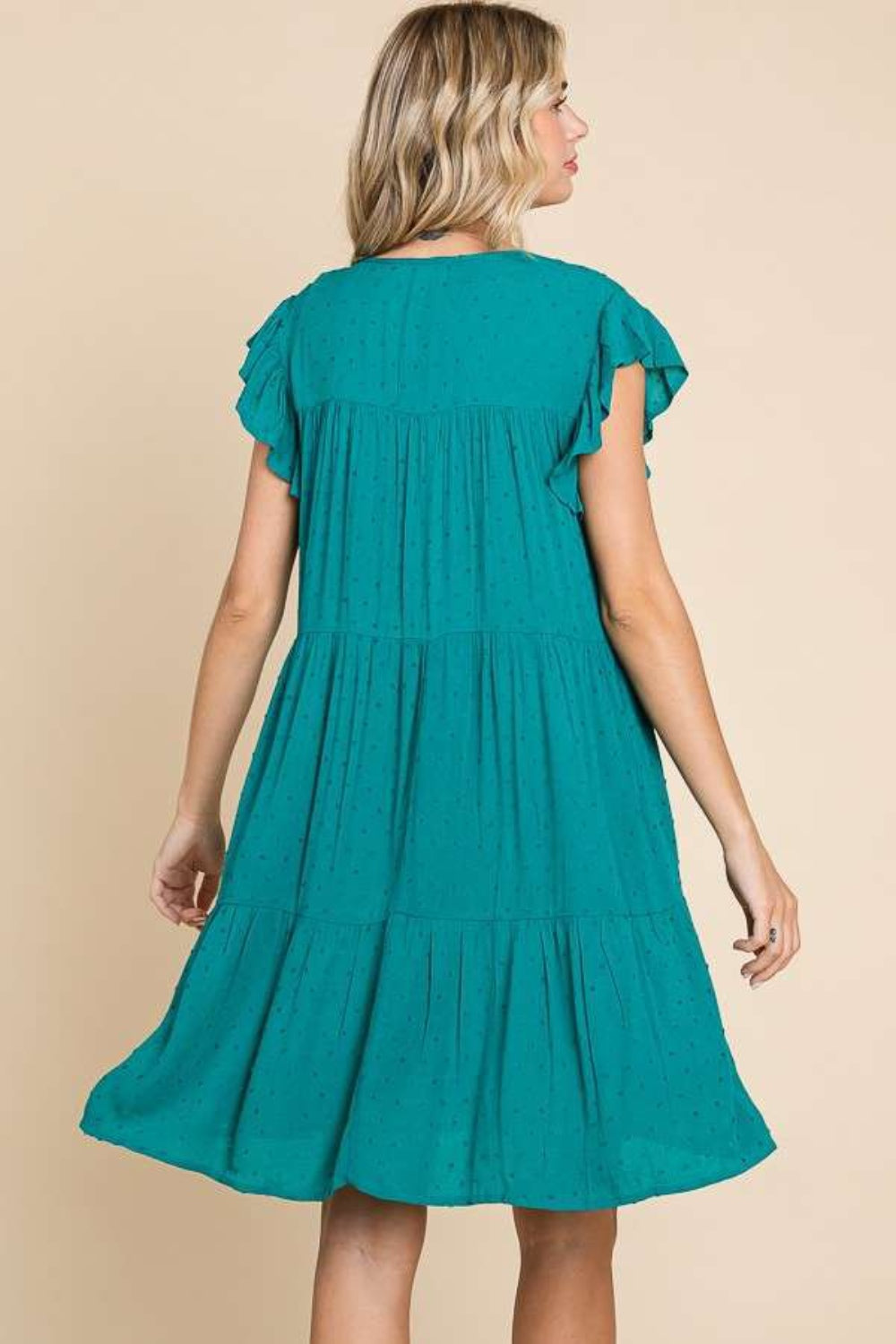 A woman stands against a beige background wearing the Culture Code Ruffle Cap Sleeve Tiered Dress, which is teal and flowing with short ruffled sleeves and a tie at the neckline. She is smiling with one hand in her pocket, showcasing the lightweight fabric.