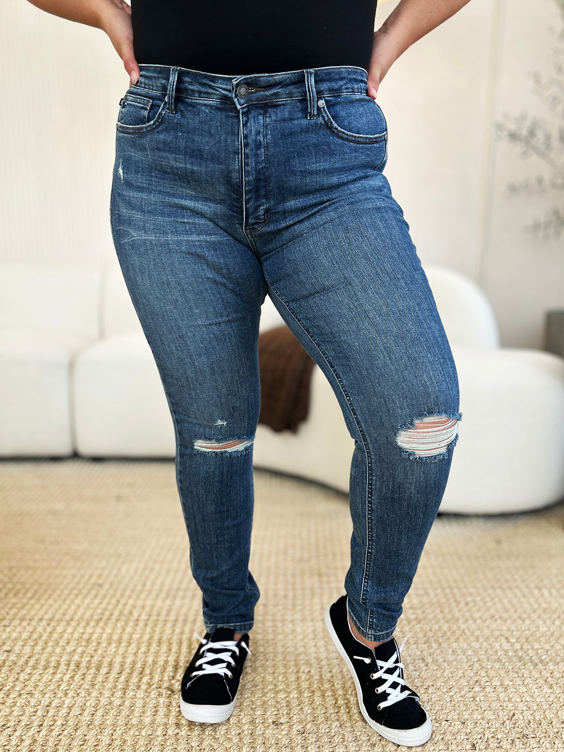 Person standing indoors wearing Judy Blue Full Size Mid Waist Distressed Slim Jeans, a black top, and black sneakers with white laces. There is a white sofa and a textured rug in the background.