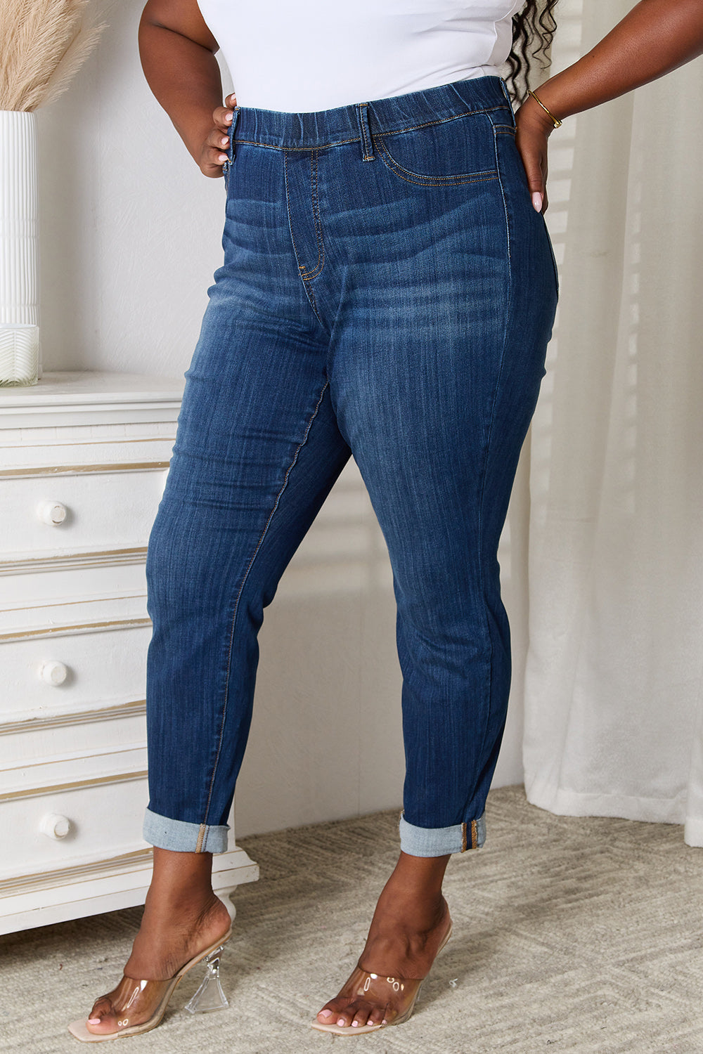 A person wearing Judy Blue Full Size Skinny Cropped Jeans with rolled-up cuffs and clear heels stands near a white dresser adorned with a decorative vase for a modern look.