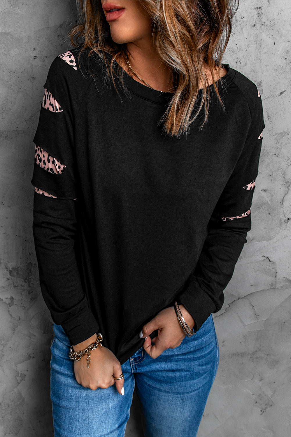 A person with wavy, highlighted hair stands facing a gray textured wall. They are wearing the Leopard Insert Black Sweatshirt with ripped leopard sleeves and blue jeans.