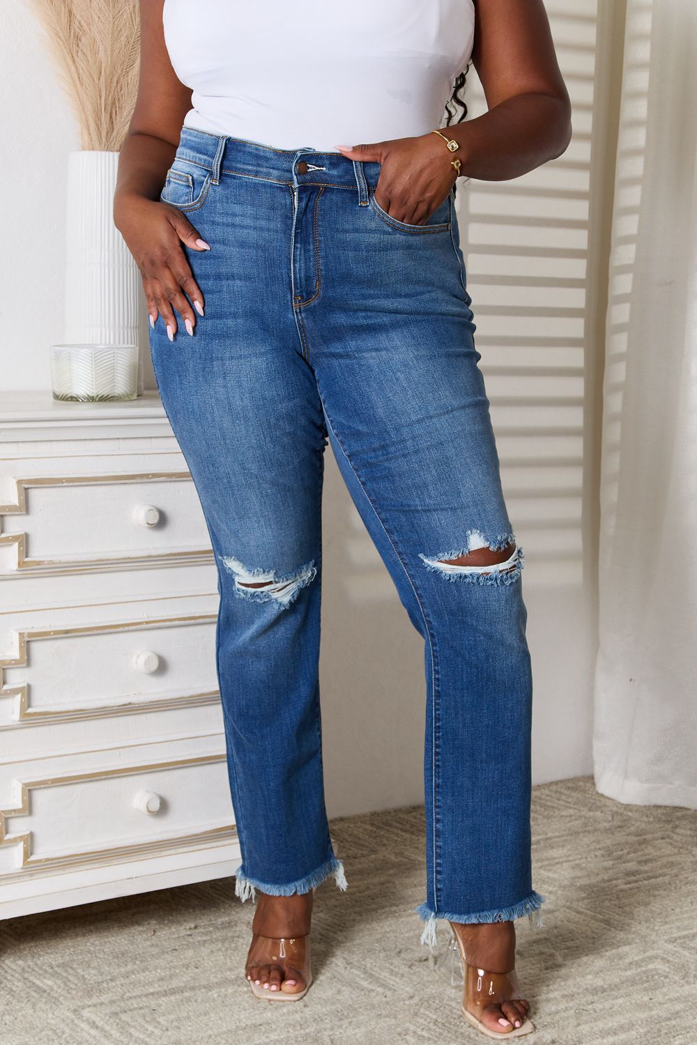 A person wearing Judy Blue Full Size Distressed Raw Hem Jeans, which are slightly stretchy and blue, along with a white top stands indoors near a white dresser.