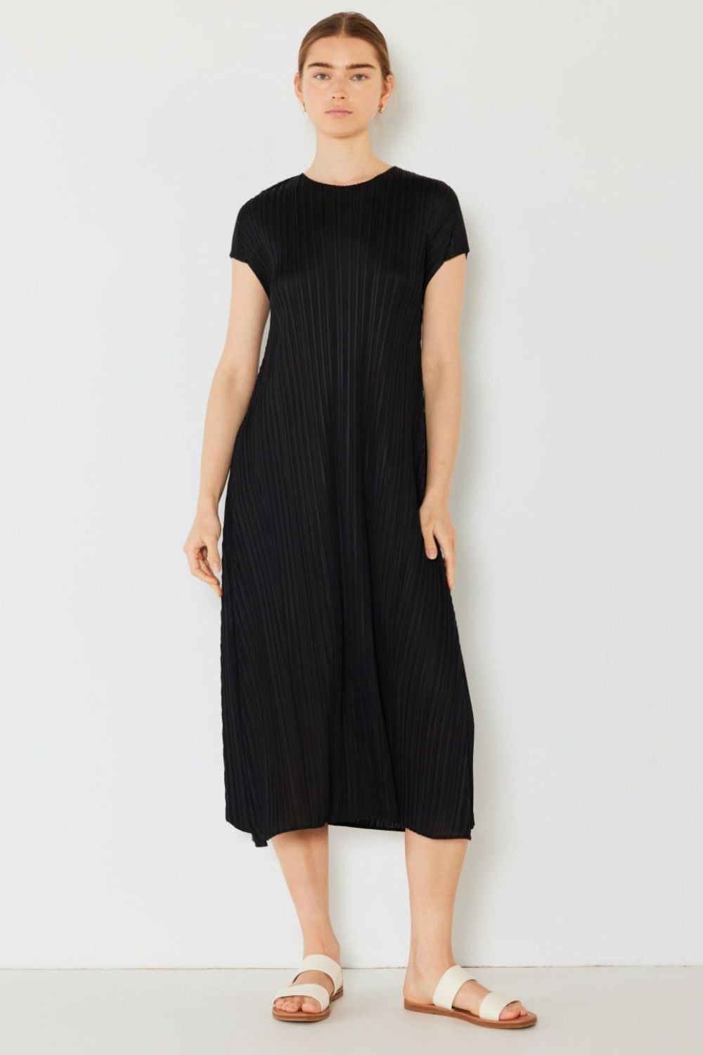 A person stands against a light wall, wearing an elegant Marina West Swim Pleated Cap Sleeve A-Line Dress in black and white sandals.