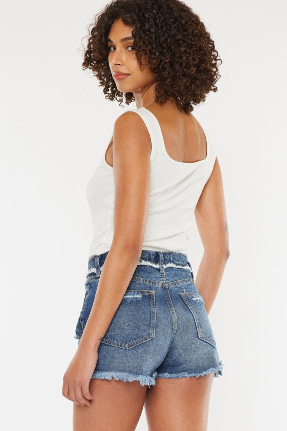 A woman with curly hair wearing a white sleeveless top and Kancan Distressed Button Fly Denim Shorts stands against a plain background, perfect for a stylish summer wardrobe.