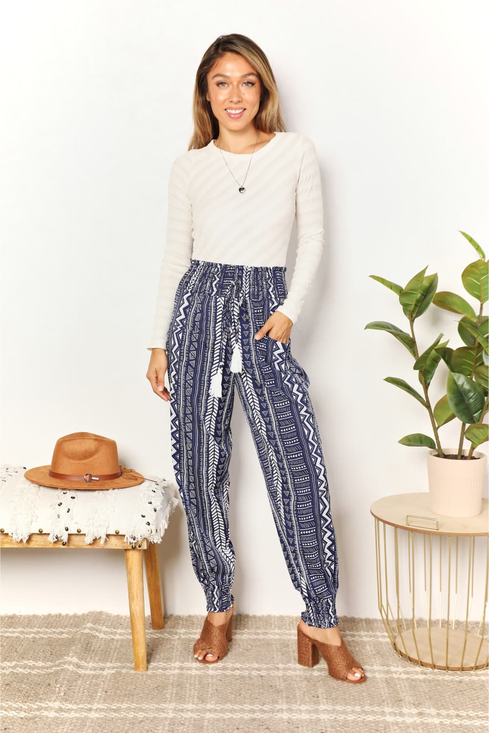 A person wearing a white long-sleeve top and Perfee Geometric Print Tassel High-Rise Pants stands next to a wooden stool and a potted plant.