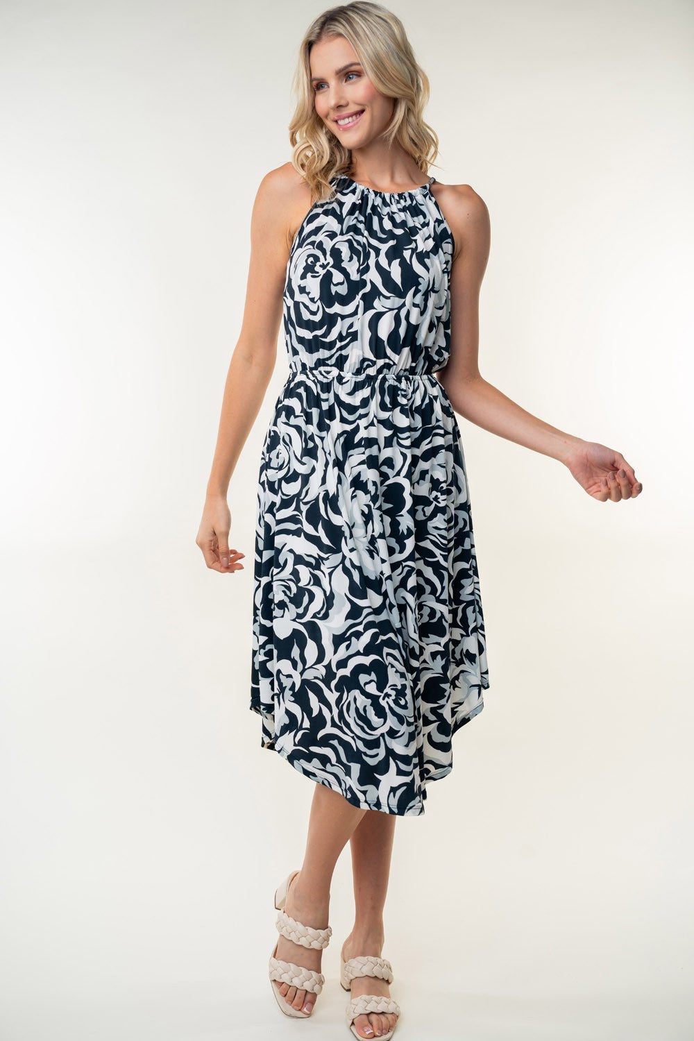 The person wearing a White Birch Tied Ruched Floral Sleeveless Knee Length Dress, featuring a black and white floral pattern, stands and smiles against a neutral background.