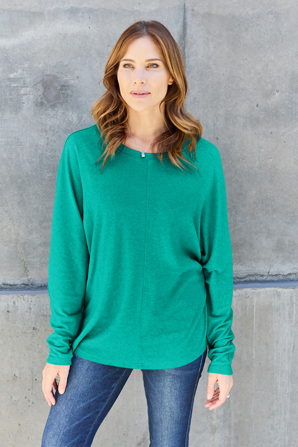 A person wearing a Double Take Full Size Round Neck Long Sleeve T-Shirt in green, blue distressed jeans with a slightly stretchy fit, and a crossbody bag, holding a beverage in a plastic cup.