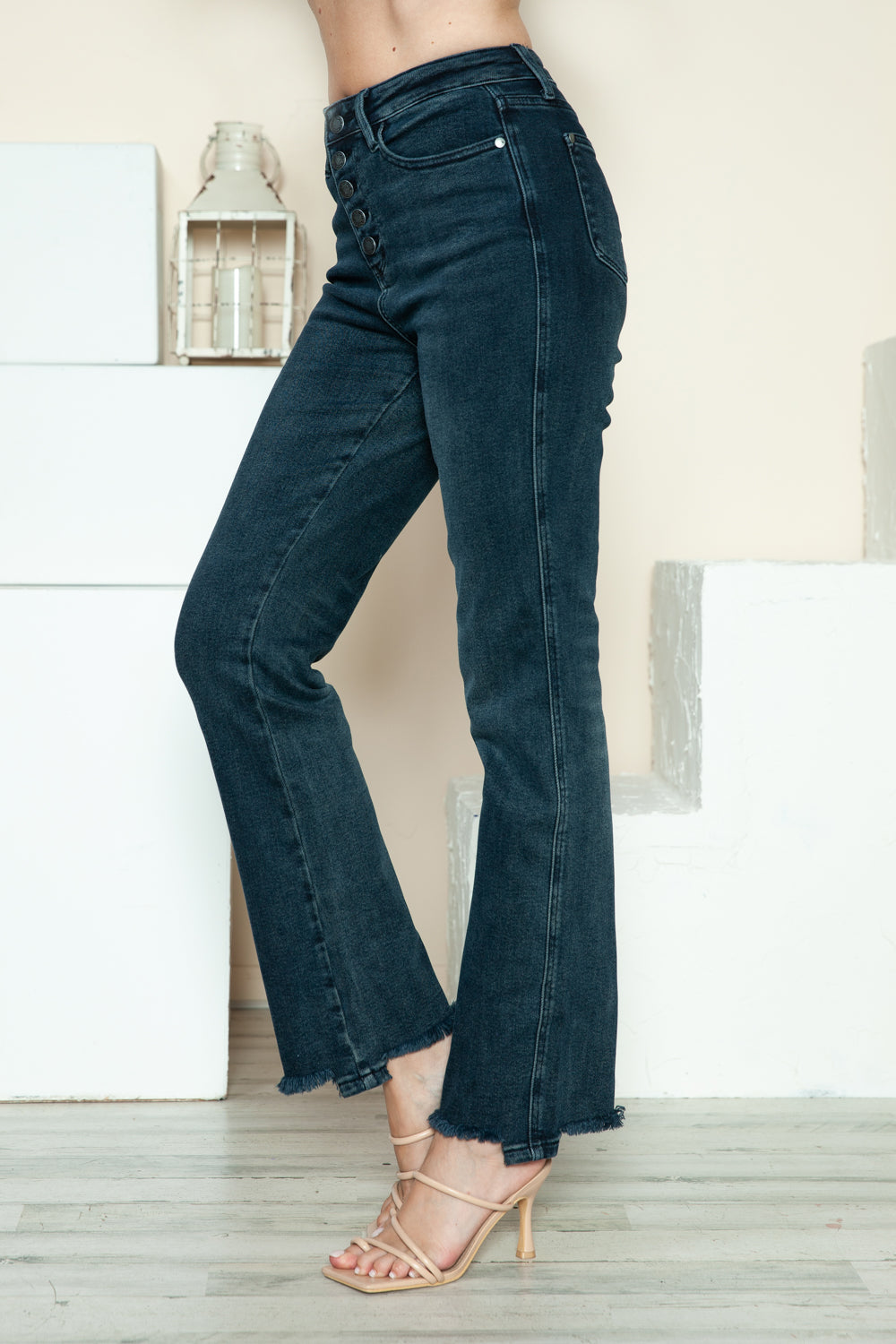 A person is modeling the Judy Blue Full Size Button Fly Hem Destroy Straight Jeans, high-waisted with distressed detailing and frayed hems, paired with light-colored open-toe heels. The background includes white blocks and a light-colored wall.