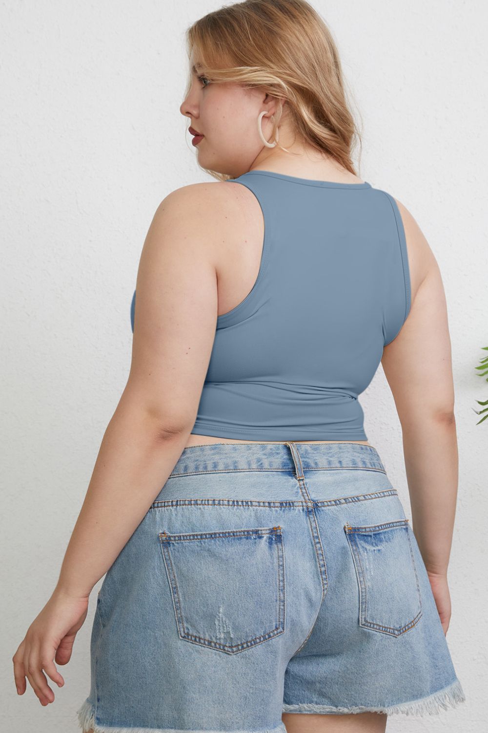 A woman with light hair wears the Basic Bae Full Size Round Neck Slim Tank in black and blue jeans, standing against a light-colored curtain.