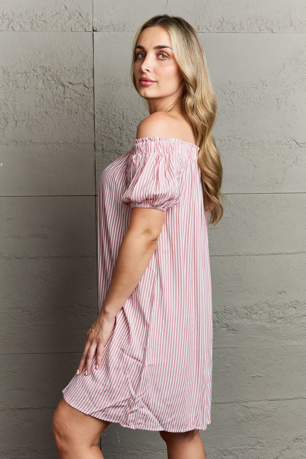 A woman exudes sophisticated elegance in the Ninexis Show Compassion Off The Shoulder Mini Dress, a pink and white striped ensemble, as she stands against a gray textured wall.