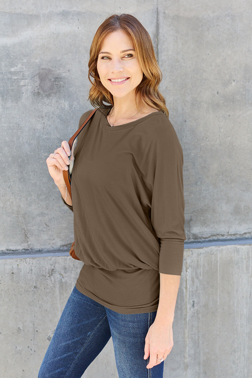 A woman in a slightly stretchy, brown Basic Bae Full Size Round Neck Batwing Sleeve top and blue jeans stands against a concrete wall, holding a brown handbag.