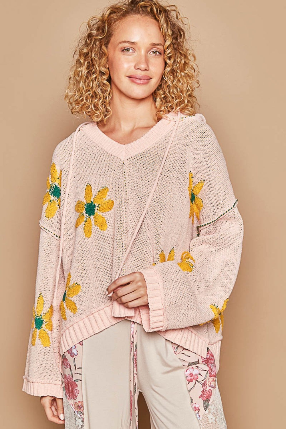 A person with curly blonde hair wears a stylish POL Floral Pattern Hooded High-Low Sweater featuring yellow flower designs and light-colored floral pants. The background is light beige.