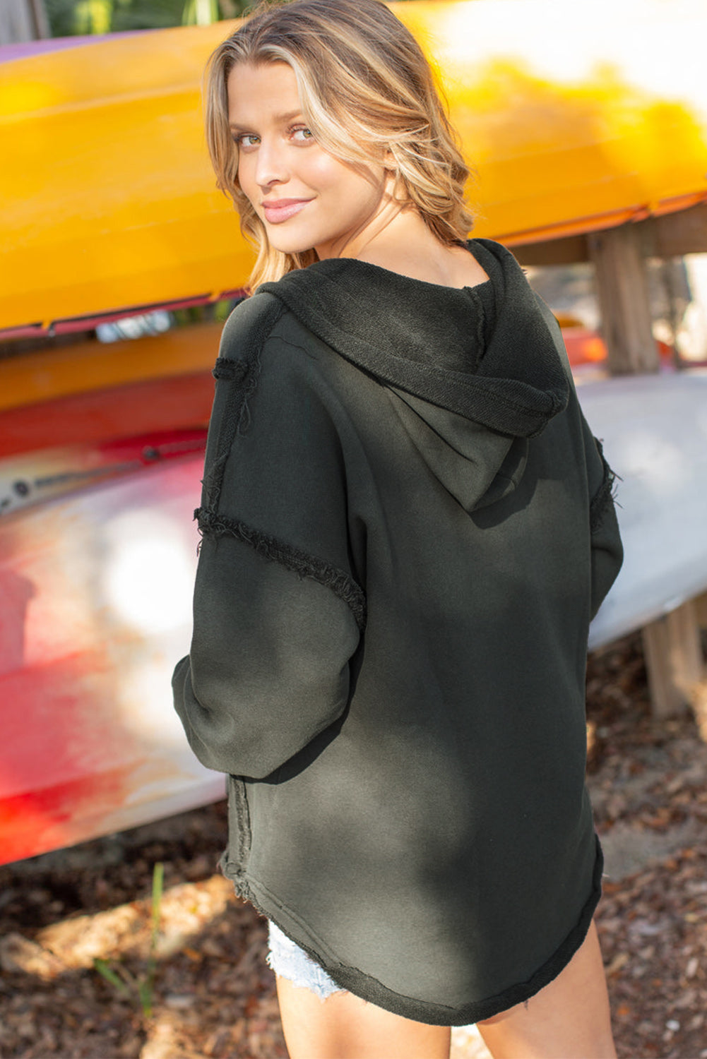 A person with blonde hair wearing a Gray Exposed Seam Raw Edge Hoodie with Pockets and denim shorts stands outdoors, looking over their shoulder. Colorful canoes are stacked in the background.