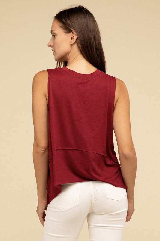 Wearing the Shark Bite Side Slit Short Sleeveless Top in maroon paired with white pants, a model is set against a beige background, exemplifying an effortlessly chic and casual wardrobe.