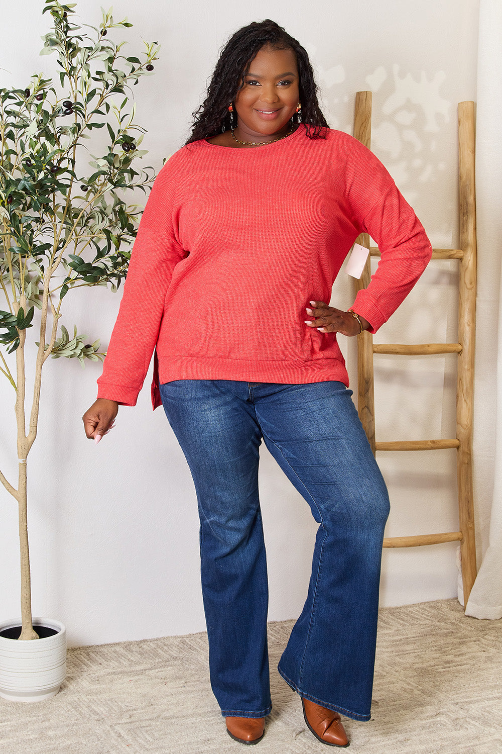 A woman wearing the Basic Bae Round Neck Drop Shoulder Slit Sweatshirt in red and blue jeans stands indoors, smiling and touching her hair, with a decorative plant and wooden ladder in the background.