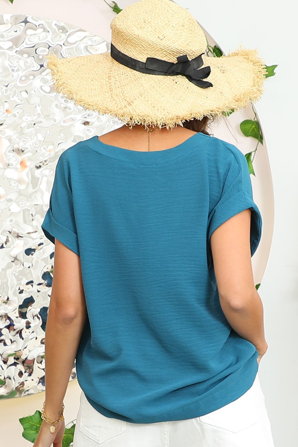 A woman wearing a stylish Ninexis V-Neck Trim Rolled Short Sleeve Shirt in light blue and high-waisted blue jeans, accessorized with hoop earrings, a necklace, and sunglasses on her head, poses in front of a beige background.