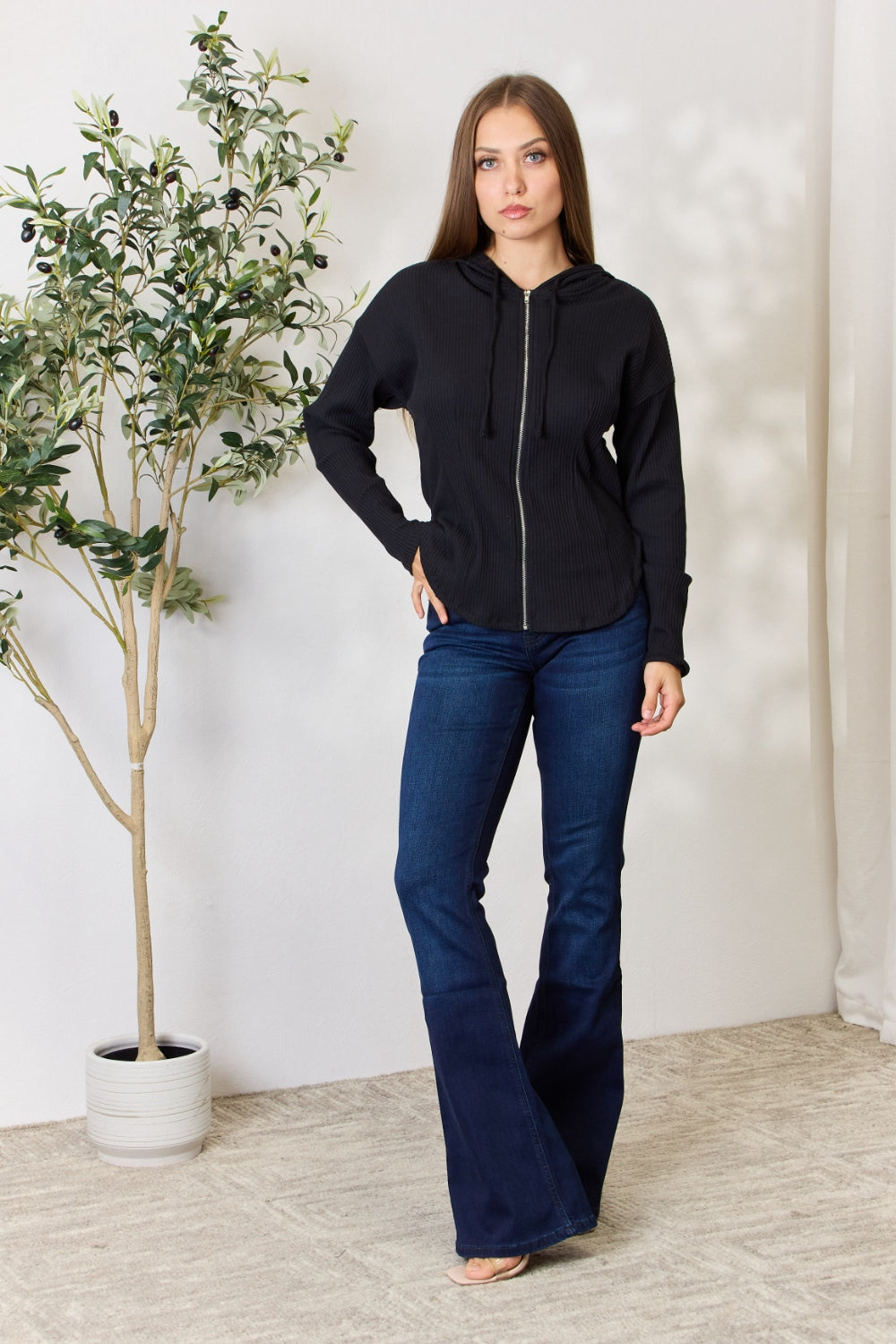 A person wearing the Culture Code Full Size Ribbed Zip Up Drawstring Hooded Jacket and blue jeans stands in front of a plain background with a small leafy plant to the left.