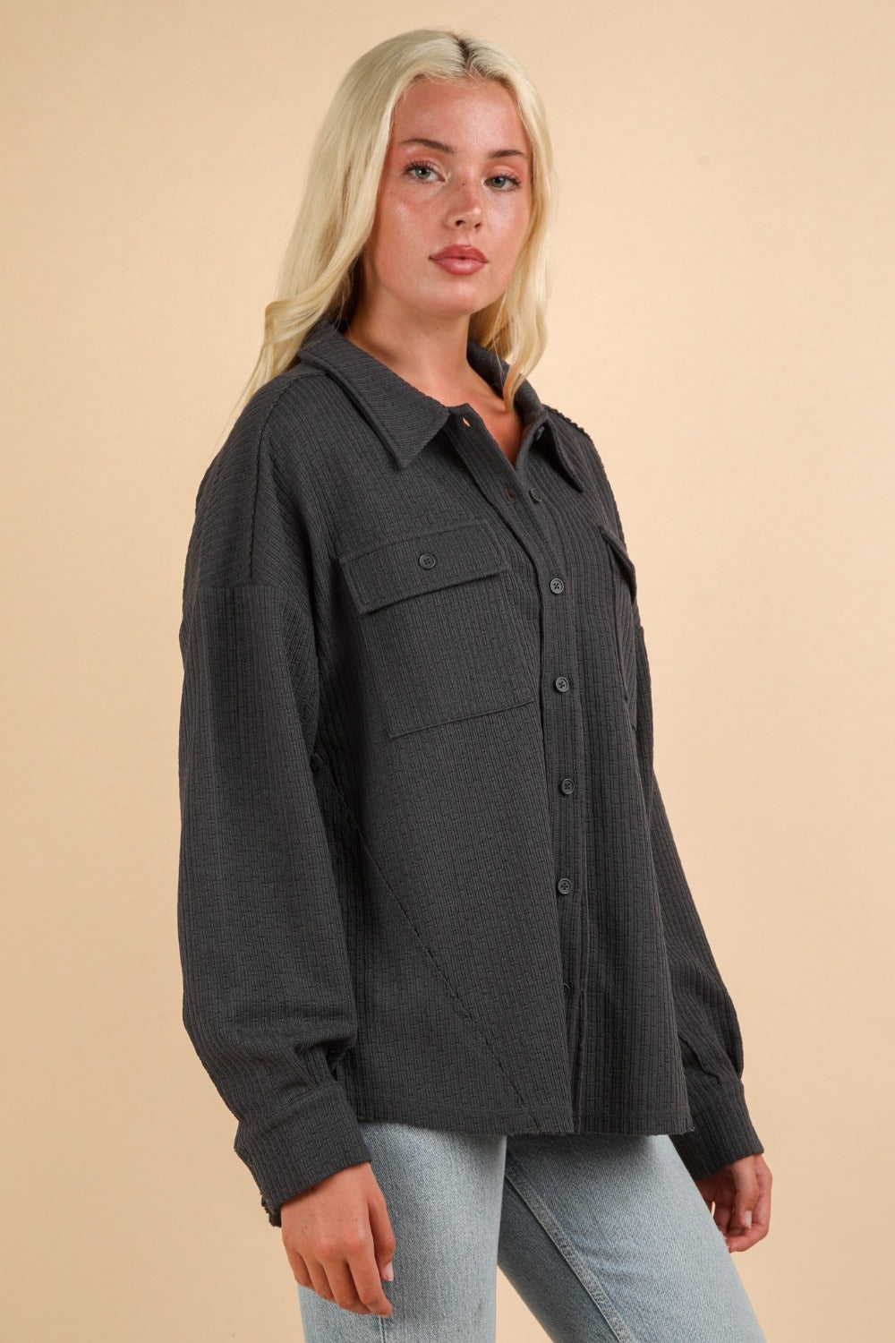 A woman with long blonde hair wears the VERY J Button Down Textured Knit Shacket in dark gray, featuring chest pockets, paired with light blue jeans. She stands against a plain beige background.