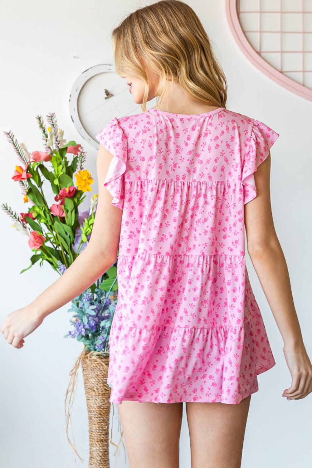 A person wearing the Heimish Full Size Floral Ruffled Tiered Top in pink paired with denim shorts is smiling while standing near a window adorned with vines, radiating a feminine and romantic vibe.