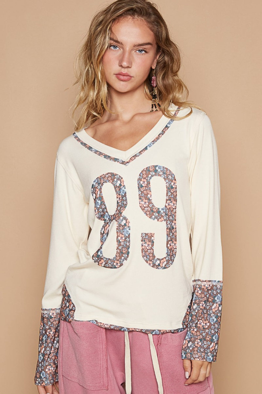 A young woman with blonde hair wears a POL V-Neck Long Sleeve Floral Number Patch T-Shirt, adorned with the number 89 in a floral design on the front and matching floral patterns on the cuffs. She also wears pink pants.