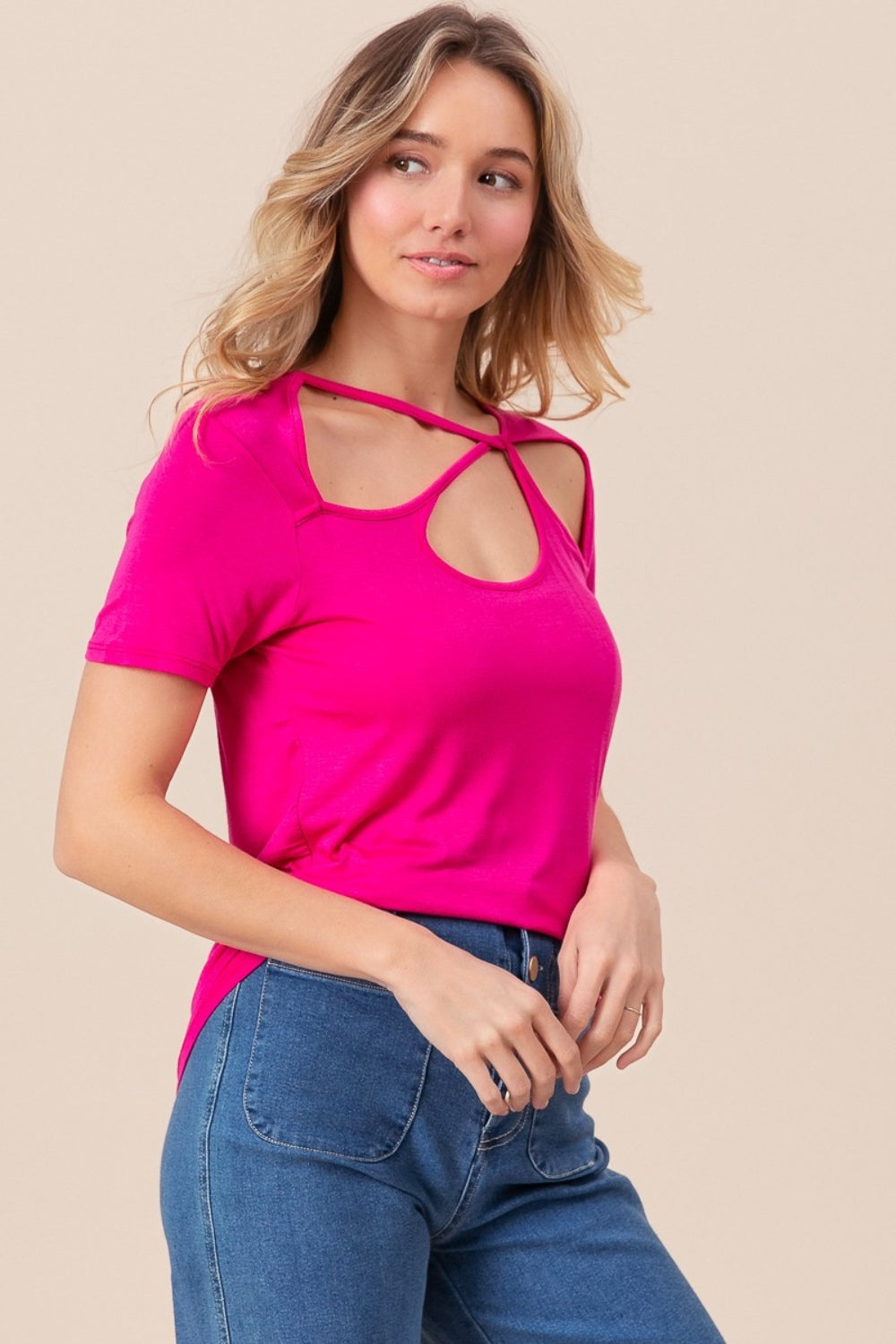 A person wearing a BiBi Cutout Asymmetrical Neck Short Sleeve T-Shirt in bright pink, along with sunglasses and denim shorts, is sitting near a wicker chair, posing confidently.