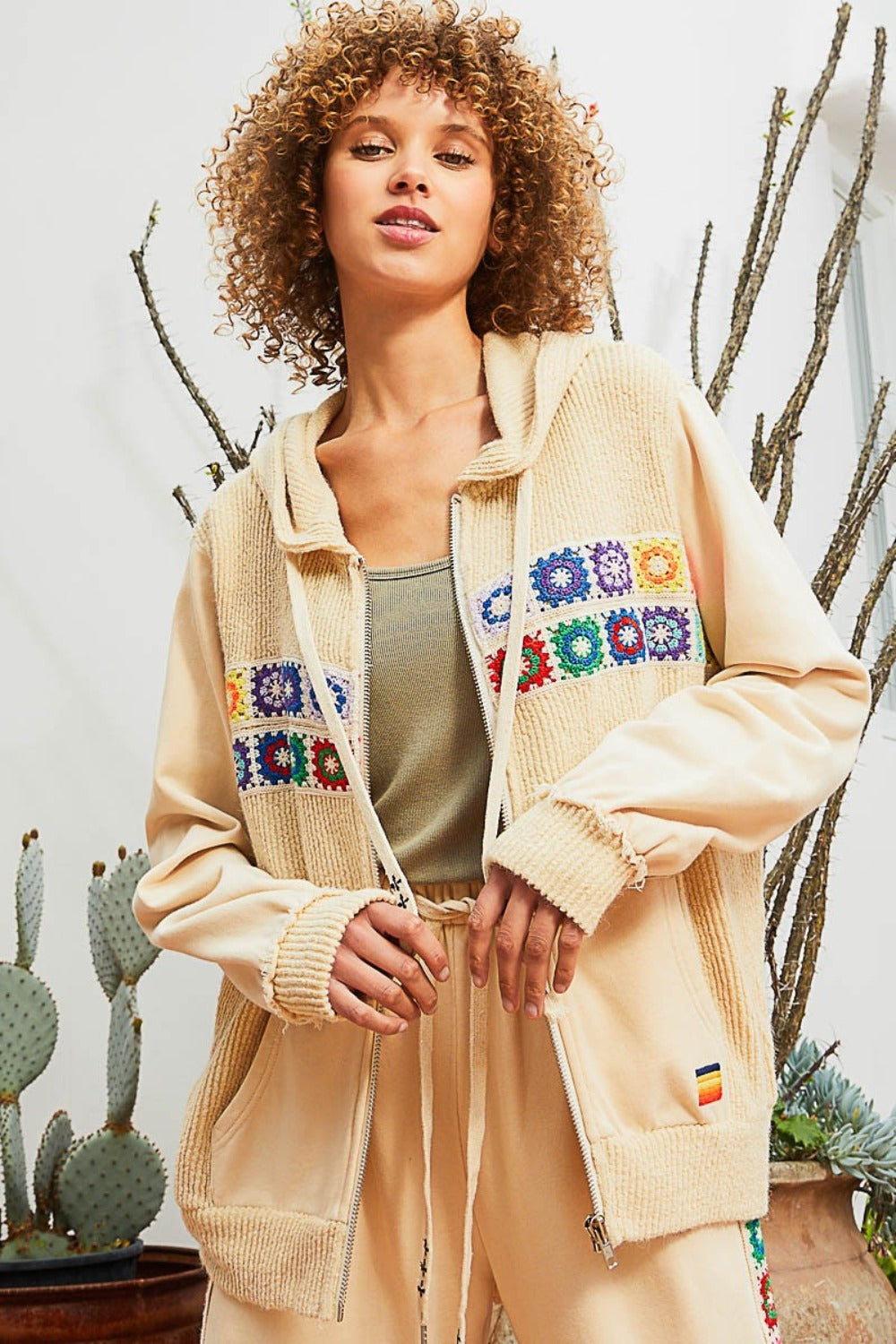 A person with curly hair stands in front of potted cactus plants, wearing the POL Contrast Square Patch Pattern Hooded Zipper Jacket. They pair it with beige pants and a green top underneath, creating a stunning look that showcases fashionable outerwear.