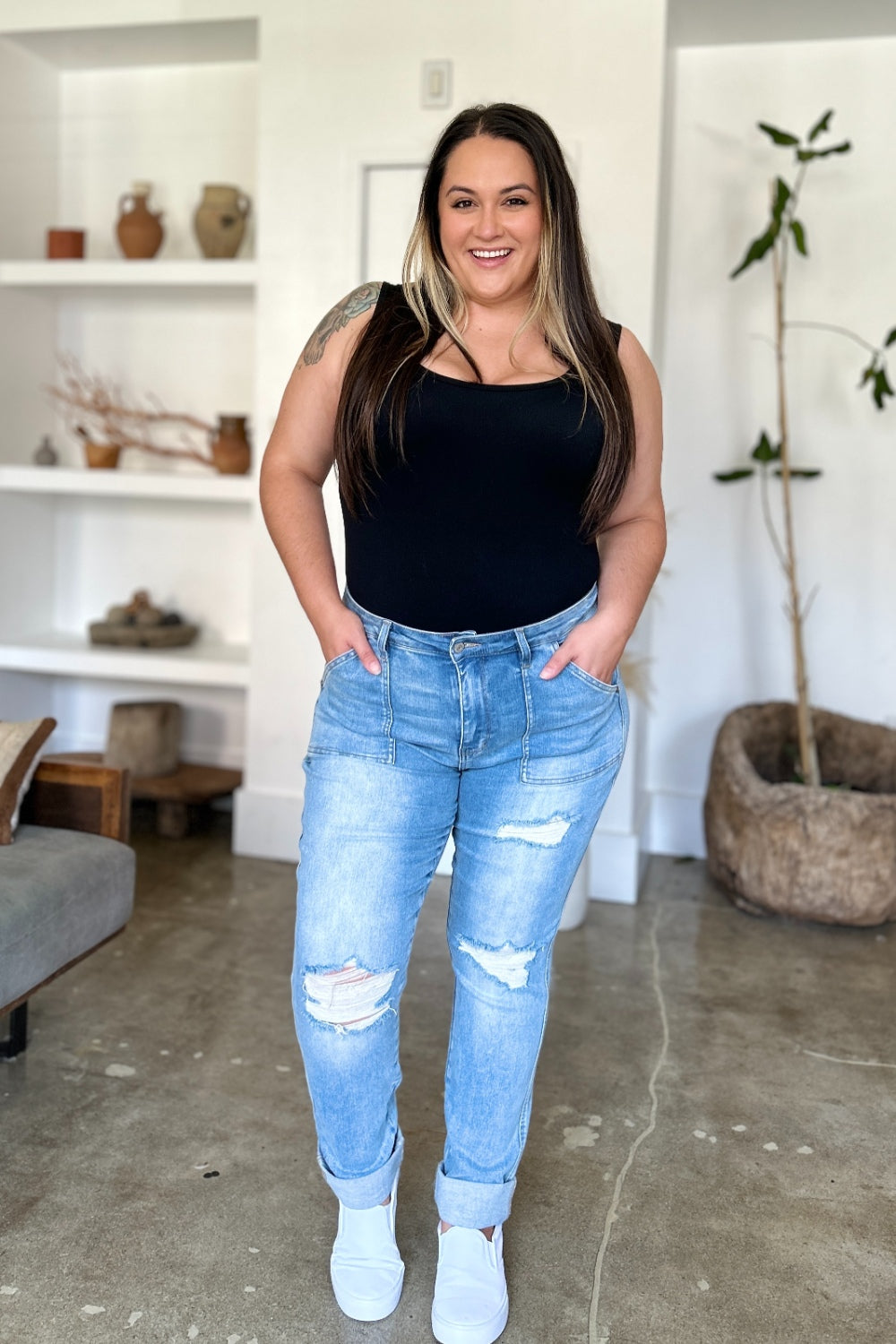 Two women are standing side by side, both clad in black and white tank tops and Judy Blue Full Size Distressed Straight Jeans with Patch Pockets, smiling at the camera in a modern living room.