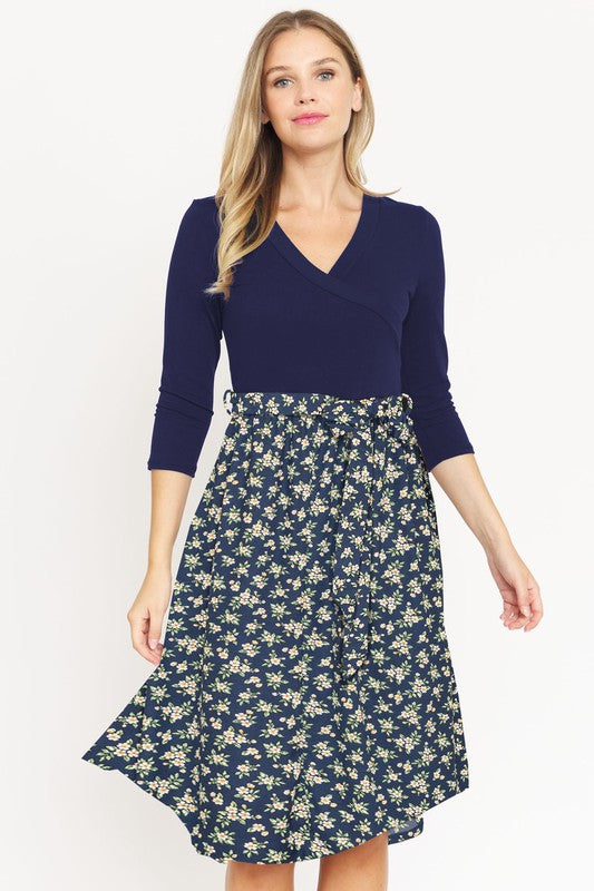 A woman in a Surplice Floral Sash Midi Dress, comprised of 95% Polyester and 5% Spandex, stands against a plain white backdrop. The knee-length dress, designed in navy blue with a floral skirt and wrapped bodice, showcases elegant craftsmanship from the United States.