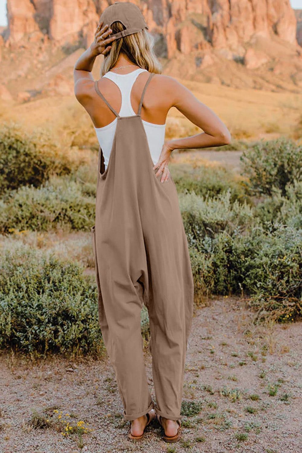 Person wearing a Double Take Full Size V-Neck Sleeveless Jumpsuit with Pockets and sandals, walking outdoors on a dirt trail with bushes in the background.