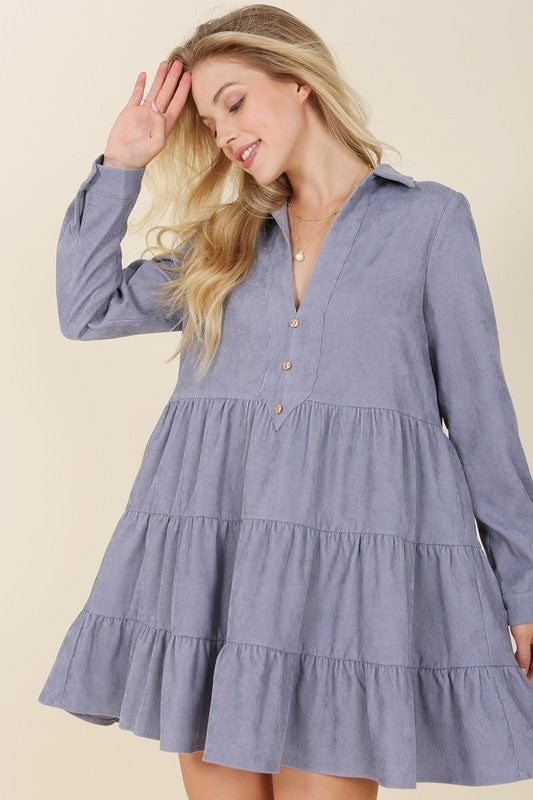 The woman in the Corduroy tiered dress, featuring a deep V-neck, smiles as she gently touches her forehead, showcasing the light blue fabric cascading elegantly.