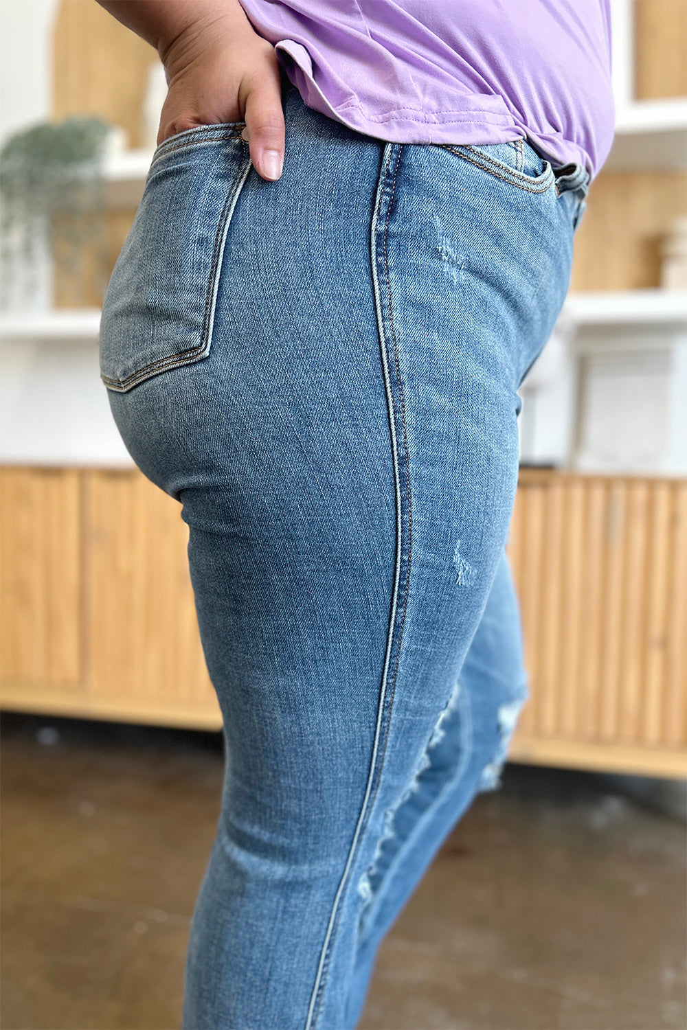 A person wearing Judy Blue Full Size Distressed Raw Hem Bootcut Jeans and a light purple top stands on a polished floor with a wooden cabinet in the background.