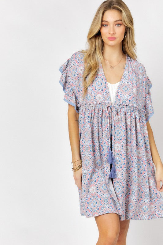 A woman stands against a white background wearing the Printed Short Sleeve Ruffle Kimono over a white top and denim shorts, effortlessly blending style and comfort.