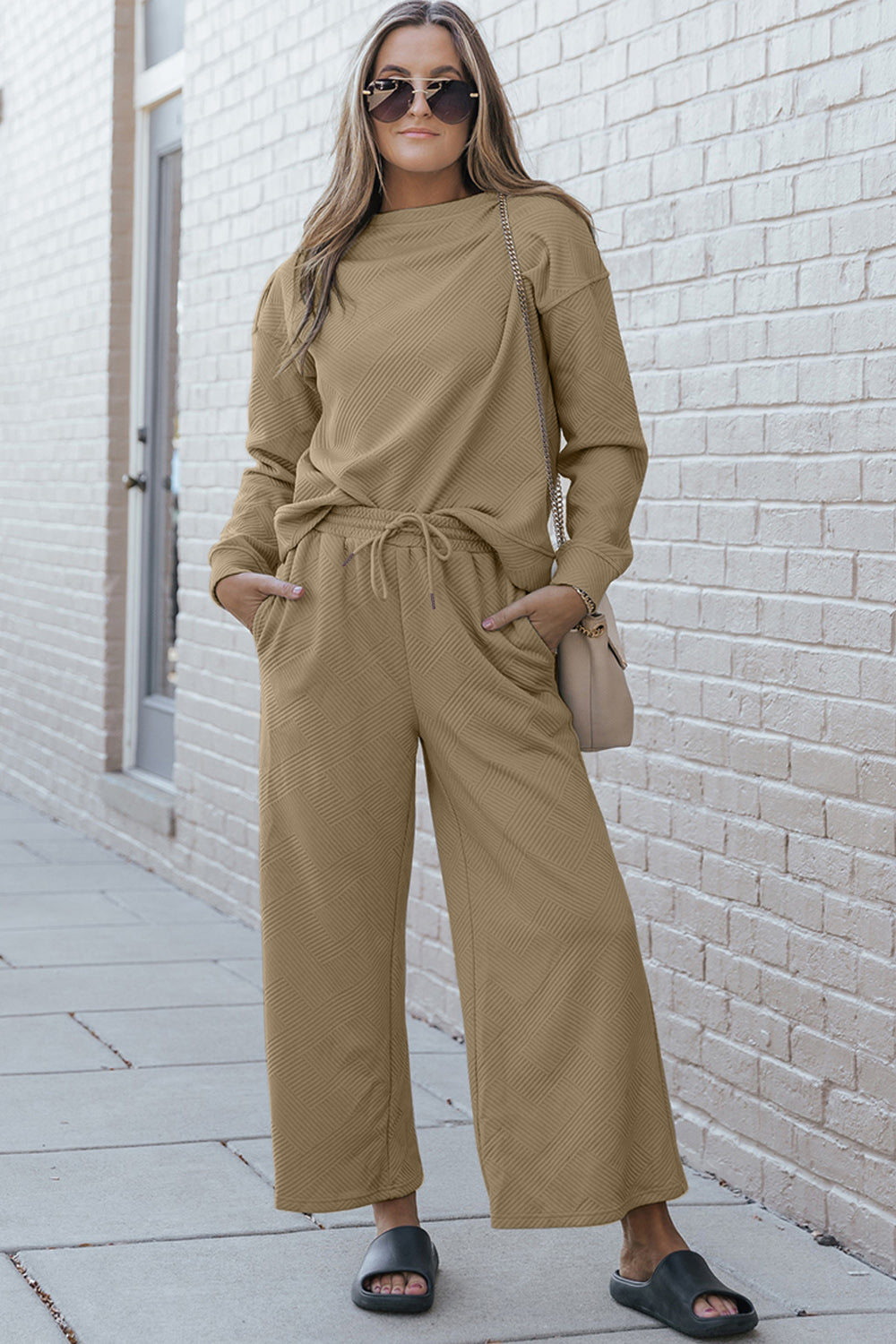 A woman with blonde hair, dressed in the Double Take Full Size Textured Long Sleeve Top and Drawstring Pants Set featuring a stretchy black long-sleeve top and matching wide-leg trousers made of rayon spandex, stands barefoot in a room with white walls and a wooden floor.