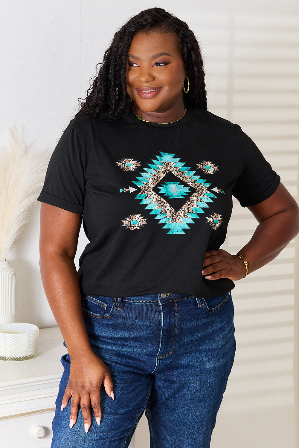 A woman stands indoors wearing the Simply Love Graphic Short Sleeve T-Shirt, a stylish black tee featuring a striking blue and white geometric pattern. Made from comfortable fabric, she has one hand on her hip and is smiling.