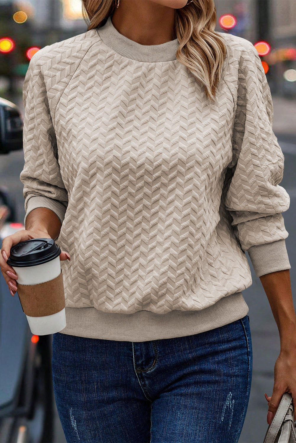 A woman in a Beige Solid Textured Raglan Sleeve Pullover Sweatshirt and blue jeans is looking to the side. She sports a brown hat and earrings, exuding a casual and comfy vibe. Standing next to dried pampas grass against a light background, she seamlessly blends with her textured sweatshirt.