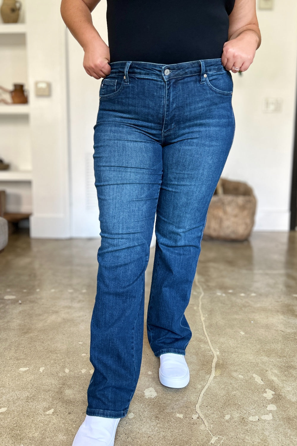 A person wearing a black top and Judy Blue Full Size Tummy Control Straight Jeans stands indoors with one hand on their hip. Their shoes are black with white soles. A plant and shelves are in the background.