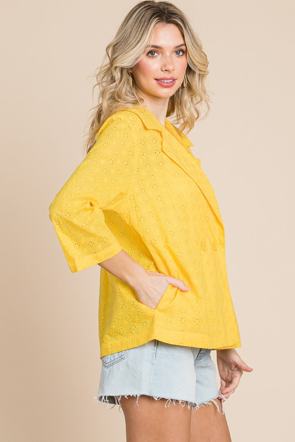 Woman in a white tank top, denim shorts, and the Culture Code Double Breasted Eyelet Jacket with Pockets in yellow, smiling against a beige background.