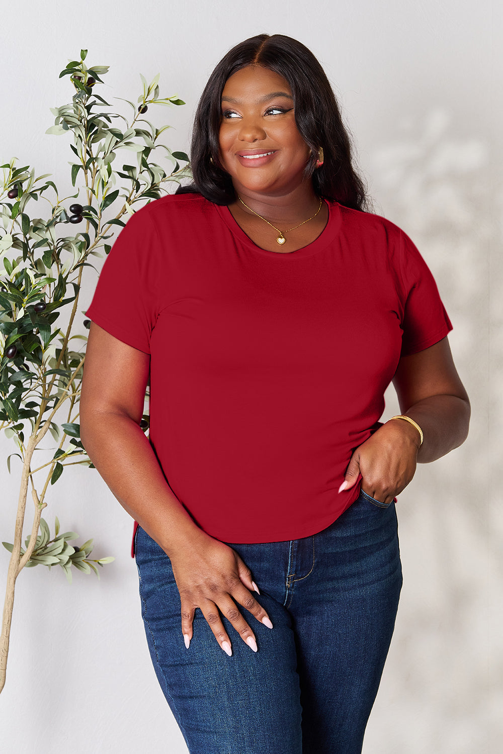 A person with wavy hair, wearing the Basic Bae Full Size Round Neck Short Sleeve T-Shirt in bright pink and beige pants, stands near a plant, smiling and looking to the side. The outfit embodies a basic style and offers slightly stretchy comfort, ideal for those who value effortless elegance.