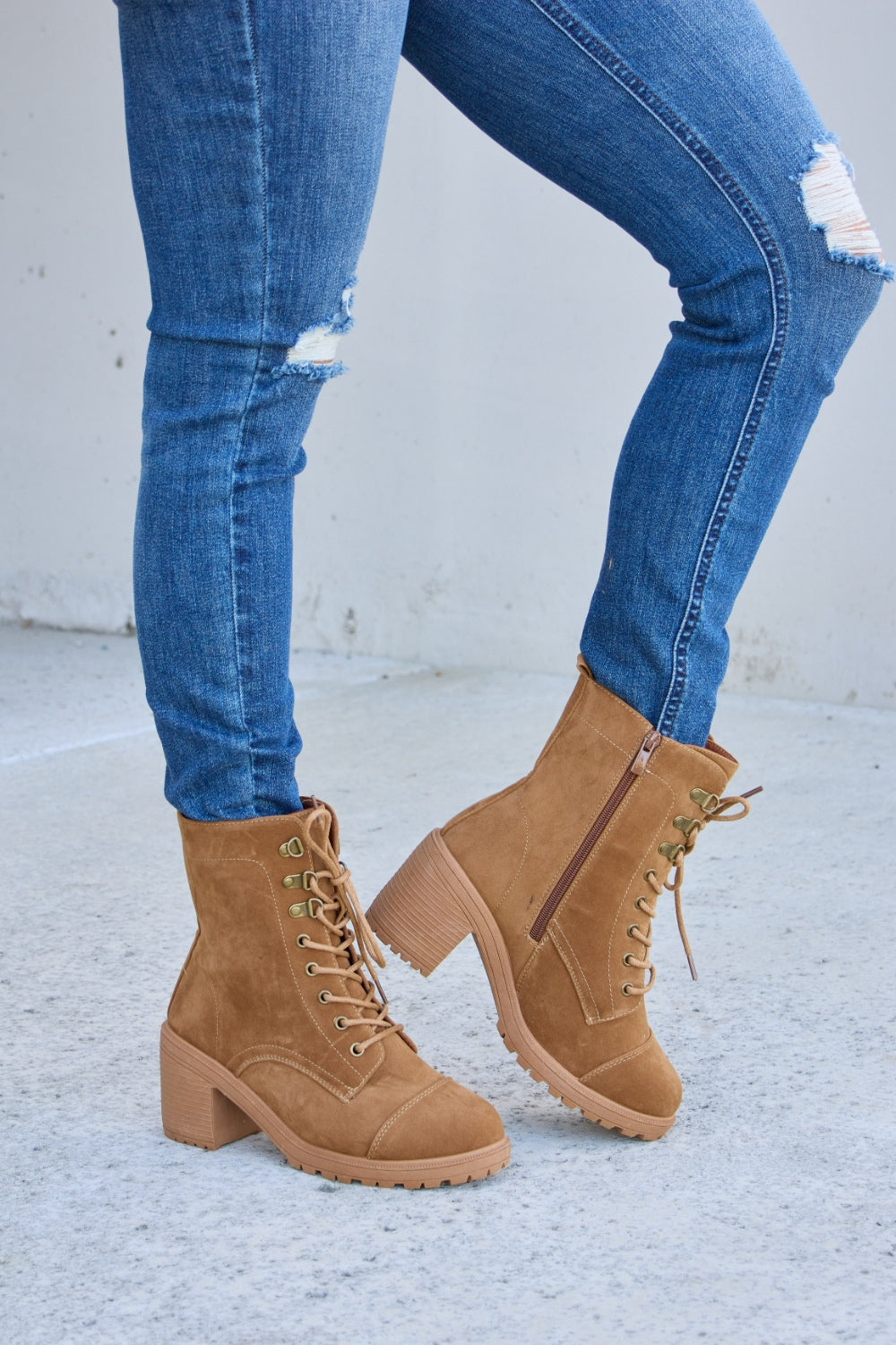 A person wearing distressed blue jeans and Forever Link Lace-Up Zipper Detail Block Heel Boots stands on a concrete surface.