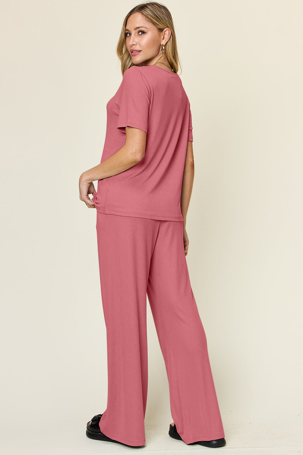 A person stands wearing the Double Take Full Size Round Neck Short Sleeve T-Shirt and Wide Leg Pants Set in a stretchy light grey fabric, with one hand in a pocket and black sandals.