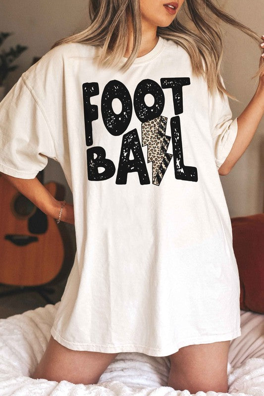 A person wearing a LEOPARD LIGHTNING FOOTBALL Graphic Tee, which features the text "FOOT BALL" and a leopard lightning football design in the center, is standing in a room with a guitar in the background.