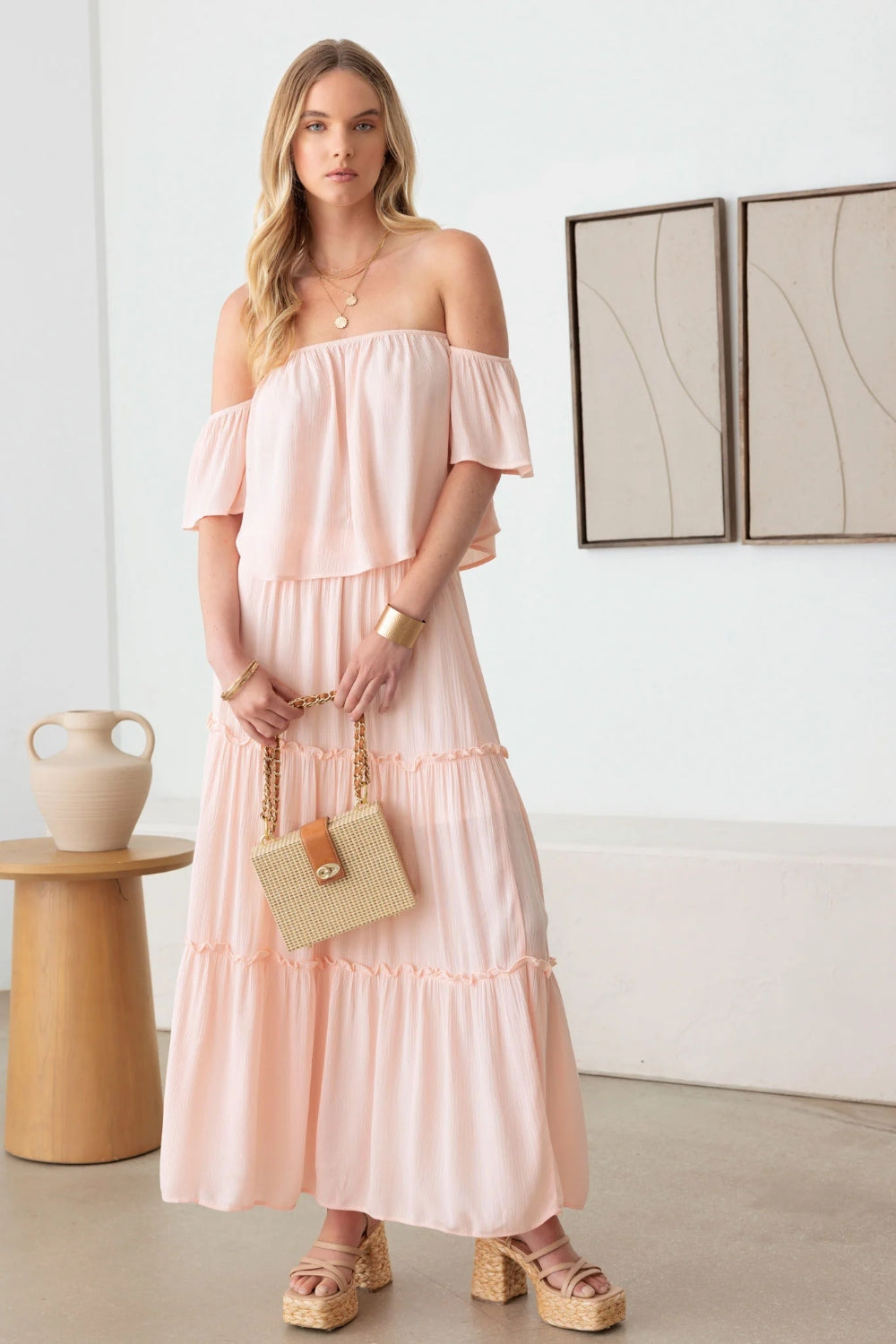 A woman in a romantic Gilli Frill Off-Shoulder Tiered Dress stands holding a beige woven handbag, with two abstract paintings on the wall behind her. She is wearing platform sandals.