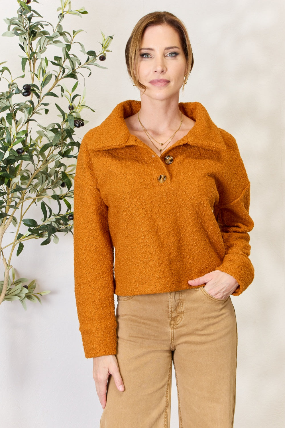 A person wears a brown textured Culture Code Full Size Half Button Turtleneck Sweatshirt made from breathable fabrics and tan pants, standing next to a leafy plant against a light background.