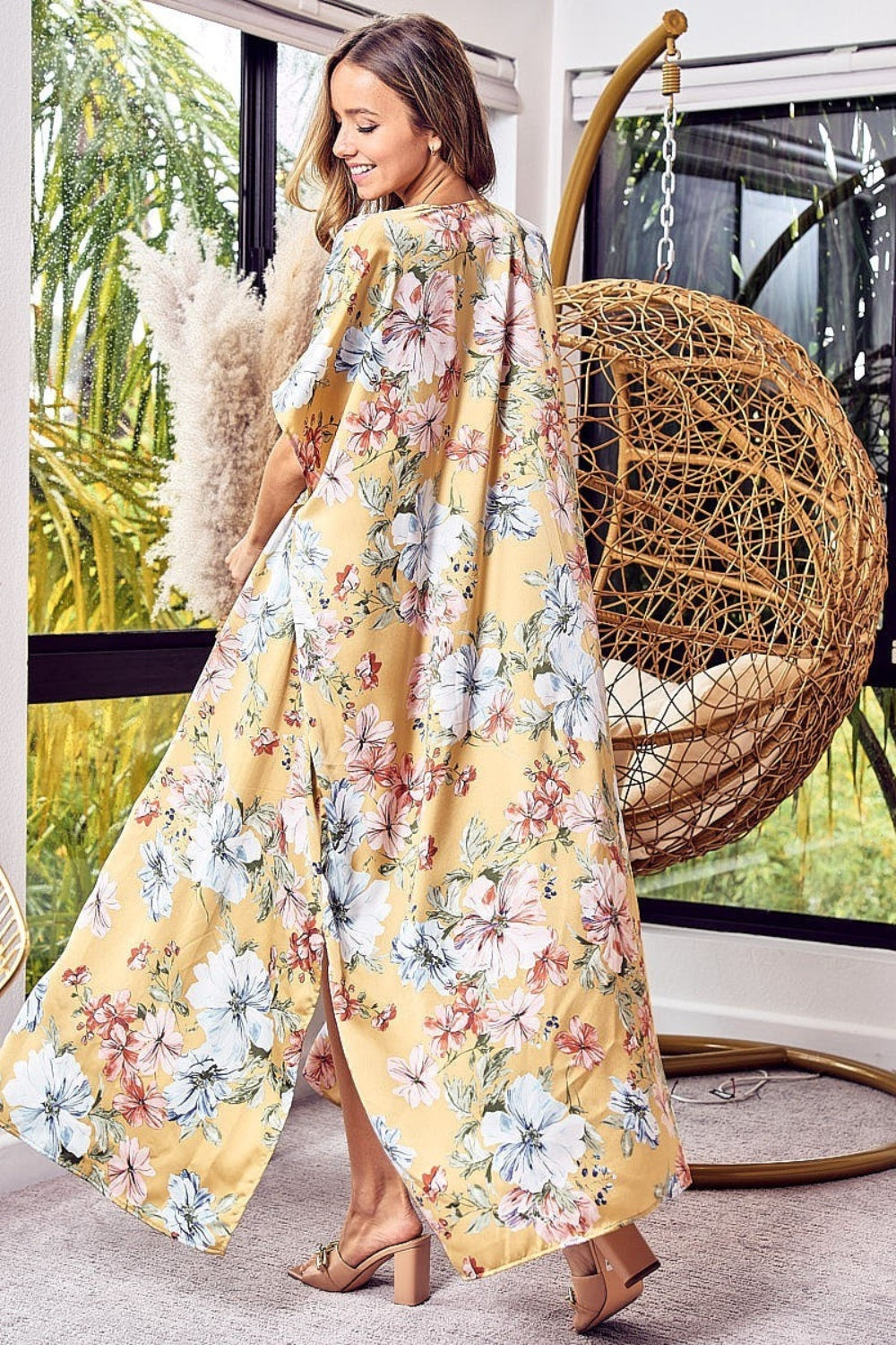 A woman dons the BiBi Floral Side Slit Longline Cardigan in yellow, paired stylishly with a white crop top and shorts. She stands indoors, holding a large sun hat with elegance. The ensemble epitomizes effortless style, echoing the vibe of a chic floral cardigan.